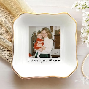 May include: A square white ceramic trinket dish with a gold-trimmed scalloped edge. The center features a printed photograph of a person holding a baby, with the black text "I love you Mom" and a small heart symbol printed below the image.