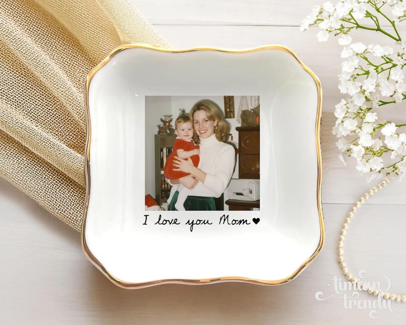 May include: A square white ceramic trinket dish with a gold-trimmed scalloped edge. The center features a printed photograph of a person holding a baby, with the black text "I love you Mom" and a small heart symbol printed below the image.
