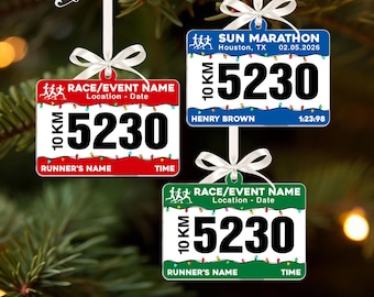 May include: Three rectangular race bib ornaments with white ribbons. One is red, one blue, and one green. Each features the text RACE/EVENT NAME, 10 KM, 5230, RUNNER'S NAME, and TIME. The blue ornament displays SUN MARATHON, HOUSTON, TX, 02.05.2026, HENRY BROWN, and 1:23:98.