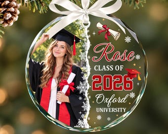 May include: A round glass ornament featuring a photo of a graduate in a black cap and gown holding a diploma. The right side displays the text Rose, Class of 2026, Oxford University, with a diploma and graduation cap icon, accented by white snowflakes and a white ribbon.