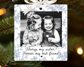May include: A square white ornament featuring a blue floral pattern and a black and white photograph of two smiling children. The text reads, Always my sister, Forever my best friend, with a small heart. A white ribbon is tied at the top.