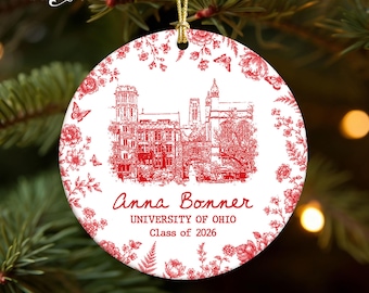 May include: A round white ceramic ornament featuring a red toile-style floral border and a detailed red illustration of a university building. The text reads Anna Bonner, UNIVERSITY OF OHIO, Class of 2026. It hangs from a gold cord.