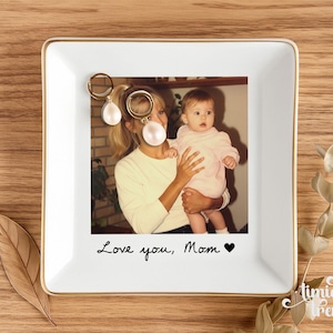May include: A square white ceramic trinket dish with a gold-toned rim featuring a printed photograph of a person holding a baby. The text Love you, Mom with a black heart symbol is printed below the photo. Two gold hoop earrings with white pearls.