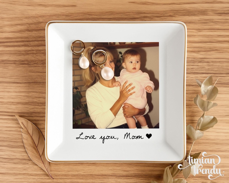 May include: A square white ceramic trinket dish with a gold-toned rim featuring a printed photograph of a person holding a baby. The text Love you, Mom with a black heart symbol is printed below the photo. Two gold hoop earrings with white pearls.