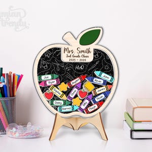 May include: Wooden apple-shaped classroom sign with a chalkboard interior, featuring student names on crayon-shaped tags. The sign includes the text "Mrs. Smith 3rd Grade Class 2025-2026 H2O". It is supported by a wooden stand.