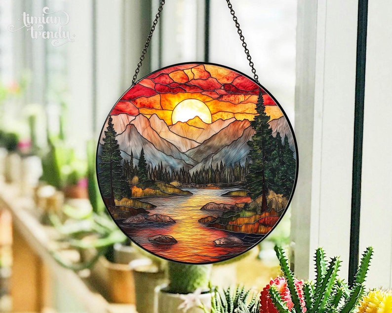 May include: A round stained glass window hanging with a chain. The window depicts a sunset over a lake and mountains with a bright orange sun and a dark blue sky.