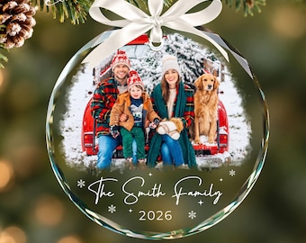 May include: A circular glass Christmas ornament featuring a photo of a family and a golden retriever sitting in the back of a red truck in the snow. The ornament has a white ribbon bow at the top and the text The Smith Family 2026 printed below.