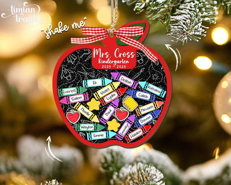 May include: A red apple-shaped Christmas ornament with a chalkboard design. It features the text "Mrs. Cross Kindergarten 2025-2026" and crayon illustrations with student names. The ornament is adorned with a red and green ribbon.