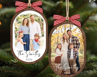 May include: Two oval-shaped wooden Christmas ornaments featuring family photographs, red and white gingham bows, and jute twine hangers. Each ornament displays the text The Smiths and the year 2026 in black script and print font on a white background.