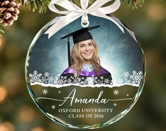May include: A round, clear glass Christmas ornament featuring a photo of a graduate in a cap and gown. The ornament is decorated with white snowflakes, a white ribbon bow, and the text Amanda, Oxford University, Class of 2026.