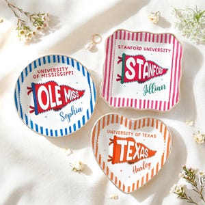May include: Three decorative ceramic dishes in different shapes: round, square, and heart. Each dish features a university pennant design with text. The round dish says "Ole Miss", the square dish says "Stanford", and the heart-shaped dish says "Texas".