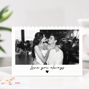 Puede incluir: Un expositor rectangular blanco con una fotograf&iacute;a en blanco y negro de dos personas abrazadas. El texto "Love you always" est&aacute; impreso en letra cursiva negra debajo de la foto, acompa&ntilde;ado de un peque&ntilde;o icono de coraz&oacute;n negro centrado.