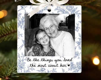May include: A square white ornament featuring a blue floral pattern and a black and white photograph of an older person and a child. The text reads, Be the things you loved the most about her. It is topped with a white satin ribbon bow.