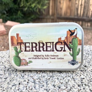 May include: A rectangular metal tin with a desert landscape illustration. The tin features the word "TERREIGN" in large letters, along with cacti, a bird, and a turtle. The design is credited to Katie Andrews and Sarah "Freedz" Conlon.