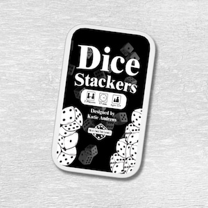 May include: A black and white game box with the title "Dice Stackers" and a design of dice. The box includes information about the game, including the number of players, play time, and age range. The game is designed by Katie Andrews.