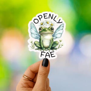 May include: A die-cut sticker featuring a green frog with translucent blue wings and a small crown of white daisy flowers on its head. The text "OPENLY FAE" is printed in black capital letters in an arc above and below the frog illustration.