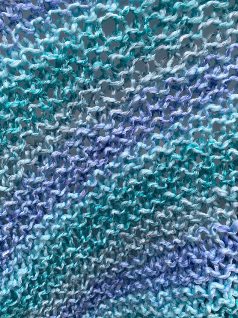 Op de afbeelding: Close-up van een gebreide stof met een gradi&euml;nt van blauwe en paarse kleuren. De stof is gemaakt van een zacht, getextureerd garen en heeft een losse, open weving.