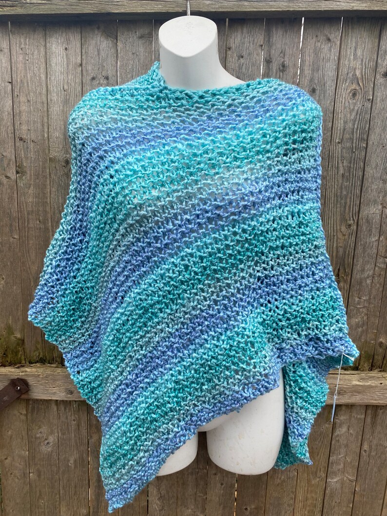 Op de afbeelding: Een blauwe en groene gebreide poncho met een losse, open structuur. De poncho is over een wit mannequin gehangen.
