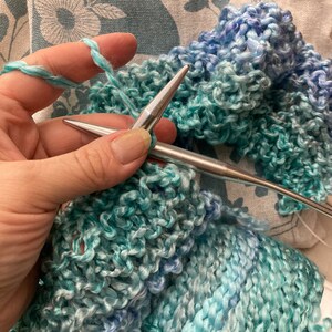 Op de afbeelding: Close-up van iemand die breien met twee zilveren breinaalden. Het garen is een lichtblauwe en turquoise ombre kleur. De bol garen bevindt zich in de rechter benedenhoek van de afbeelding. Het garenlabel vermeldt "6 oz Net Wt 185 yds."