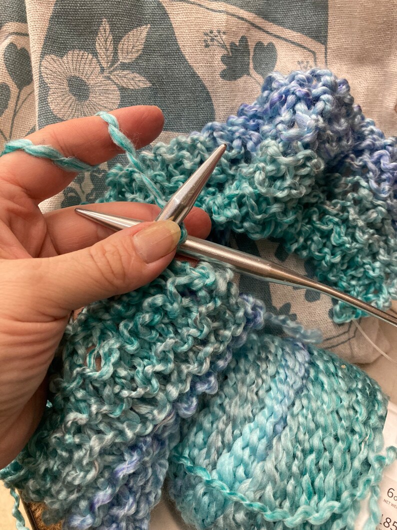 Op de afbeelding: Close-up van iemand die breien met twee zilveren breinaalden. Het garen is een lichtblauwe en turquoise ombre kleur. De bol garen bevindt zich in de rechter benedenhoek van de afbeelding. Het garenlabel vermeldt "6 oz Net Wt 185 yds."