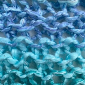 Op de afbeelding: Close-up van een gebreide stof met een gradi&euml;nt van blauwe en turquoise garen. De stof is gemaakt met een eenvoudig steekpatroon, waardoor een textuur ontstaat.