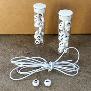 Pu&ograve; includere: Due provette di plastica trasparente piene di perline bianche con numeri e simboli neri. Sono presenti anche un cordoncino bianco e due perline sfuse con uno zero e un trattino. Gli oggetti sono su una superficie neutra.