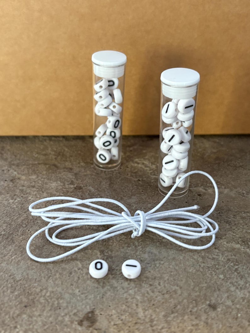 Pu&ograve; includere: Due provette di plastica trasparente piene di perline bianche con numeri e simboli neri. Sono presenti anche un cordoncino bianco e due perline sfuse con uno zero e un trattino. Gli oggetti sono su una superficie neutra.