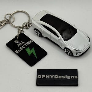 May include: A white toy car with a black roof is next to a black keychain tag with "ALL ELECTRIC" and a green lightning bolt. A small black rectangle with "DPNYDesigns" is also present. The keychain has a silver clasp.