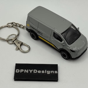 May include: A gray die-cast Transit van keychain with black wheels and a yellow stripe. The keychain includes a silver key ring and clasp. A black rectangular base with the text "DPNYDesigns" is also present.