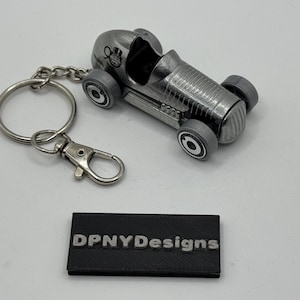 May include: A silver race car keychain with a key ring and clasp. The car has gray wheels and a detailed body. The text "DPNYDesigns" is on a black rectangular base.