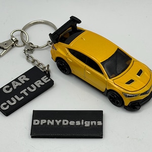 Puede incluir: Un llavero de coche de juguete amarillo con un alerón trasero negro y ruedas negras. Unido a la anilla metálica hay una etiqueta rectangular negra con el texto blanco CAR CULTURE. Una placa rectangular negra separada muestra el texto DPNYDesigns.