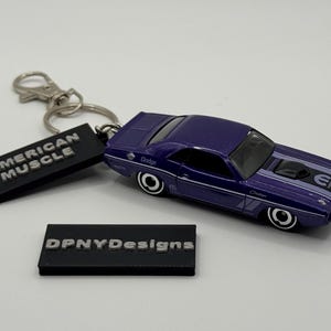 May include: A purple Dodge Challenger keychain with white racing stripes and black wheels. The keychain includes a black tag that reads "AMERICAN MUSCLE". The car has the word "Dodge" on the side.