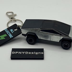 May include: A silver and black Cybertruck toy car keyring with a black tag that reads "ALL ELECTRIC" and a green lightning bolt. The keyring also has a small black rectangular block that reads "DPNYDesigns".