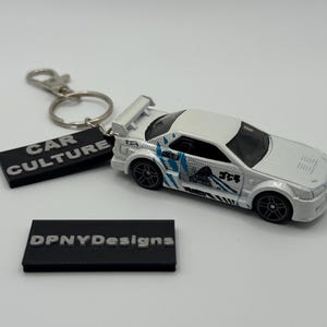 May include: A white toy car with blue and black graphics, a rear spoiler, and black wheels. The car is part of a keychain with a black tag that reads "CAR CULTURE". A second black tag reads "DPNYDesigns".