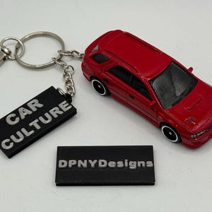 May include: A red toy car with black wheels and a keychain. The keychain has a black rectangular tag that reads "CAR CULTURE" in white letters. A second black rectangular tag with "DPNYDesigns" is also present.