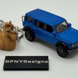 May include: A blue toy SUV and a horse-shaped keyring. The keyring is brown with a silver chain. A black rectangular block with the text "DPNYDesigns" is also present.