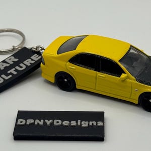 May include: A yellow toy car with a black hood and black wheels. The car is part of a keychain with a silver ring and a black rectangular tag that reads "CAR CULTURE". A second black rectangular tag reads "DPNYDesigns".