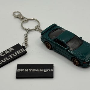 May include: A dark green toy car keychain with a silver chain and a black tag that says "CAR CULTURE". A second black tag reads "DPNYDesigns". The car has gold-colored wheels.