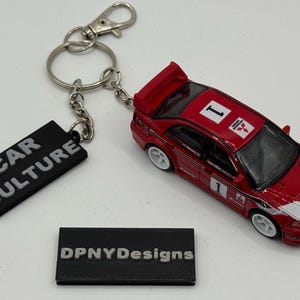 Könnte beinhalten: Ein rotes Spielzeugauto mit weißen Akzenten und der Nummer 1 auf dem Dach und der Seite, befestigt an einem Schlüsselanhänger mit einem schwarzen Anhänger mit der Aufschrift "CAR CULTURE". Ein kleines schwarzes Etikett mit "DPNYDesigns" ist ebenfalls vorhanden.
