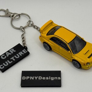 Pode incluir: Um porta-chaves de carro de brinquedo amarelo com um porta-chaves e uma corrente prateados. O porta-chaves tem uma etiqueta preta que diz "CAR CULTURE". Uma segunda etiqueta preta diz "DPNYDesigns".