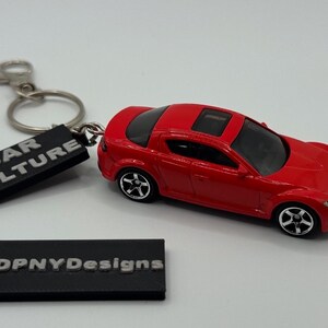 Puede incluir: Un coche de juguete rojo con ruedas negras y techo solar está junto a un llavero con las palabras "CAR CULTURE" en un rectángulo negro. También hay un segundo rectángulo negro con "DPNYDesigns".