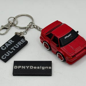 May include: A red toy car keychain featuring a stylized sports car with white-rimmed wheels and a windshield decal reading CAR TUNED. Attached is a black rectangular tag with the text CAR CULTURE and a separate black tag labeled DPNYDesigns.
