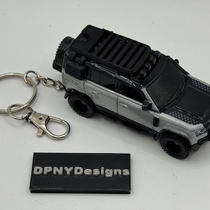 May include: A silver and black miniature SUV keychain with a black roof rack and spare tire. The keychain includes a silver key ring and clasp. A black rectangular tag reads "DPNYDesigns".