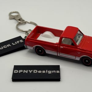 Puede incluir: Un llavero de camioneta roja de juguete con una caja blanca y detalles plateados. El llavero incluye una etiqueta negra con el texto "TRUCK LIFE" y otra con "DPNYDesigns". El camión tiene ruedas negras.