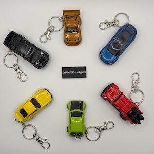 Puede incluir: Una colecci&oacute;n de llaveros de coches de juguete en varios colores, incluyendo negro, dorado, azul, amarillo, verde y rojo. Cada coche est&aacute; unido a un llavero plateado con un cierre de mosquet&oacute;n. La imagen incluye un rect&aacute;ngulo negro con el texto "DPNYdesigns".