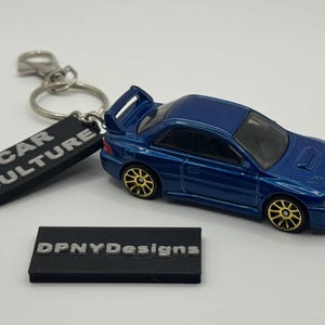 May include: A blue die-cast model car with gold-colored wheels, attached to a keychain with a black tag that reads "CAR CULTURE". A second black tag reads "DPNYDesigns".