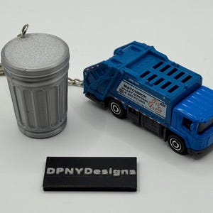 May include: A blue Matchbox garbage truck toy next to a silver trash can keychain. The truck has black wheels and the words "Valley Disposal & Recycling" printed on the side. A black rectangular sign reads "DPNYDesigns".