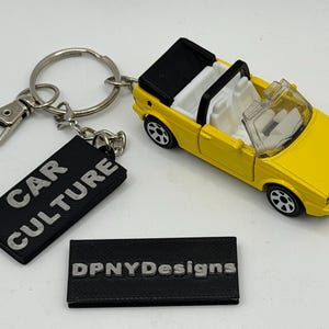 Puede incluir: Un llavero de coche de juguete descapotable amarillo con techo negro e interior blanco. Unido a la anilla metálica hay una etiqueta rectangular negra con el texto blanco CAR CULTURE. Una etiqueta negra separada muestra el texto blanco DPNYDesigns.