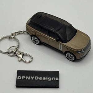May include: A gold-colored toy car keychain with a black roof and silver wheels. The keychain includes a silver key ring and clasp. A black rectangular block with the text "DPNYDesigns" is also present.