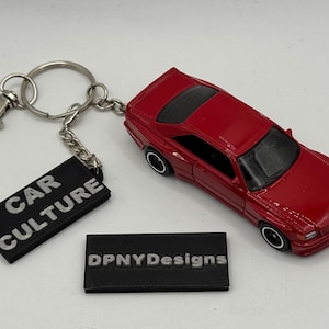 May include: A red die-cast car keychain with a silver keyring and chain. The keychain includes a black rectangular tag that reads "CAR CULTURE". A second black tag reads "DPNYDesigns".